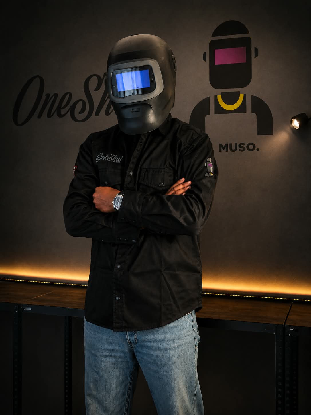 MUSO con la OneShot FR Jacket OFFICINA 01 e il casco da saldatura, a braccia incrociate davanti al logo OneShot in officina